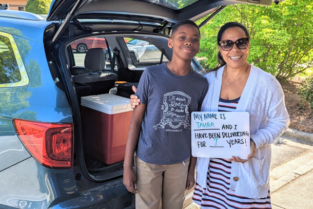 Woman volunteer and her son standing in front of a cooler in the back of their car holding up a dry erase board that says, My name is Tahra and I have been delivering for 7 years